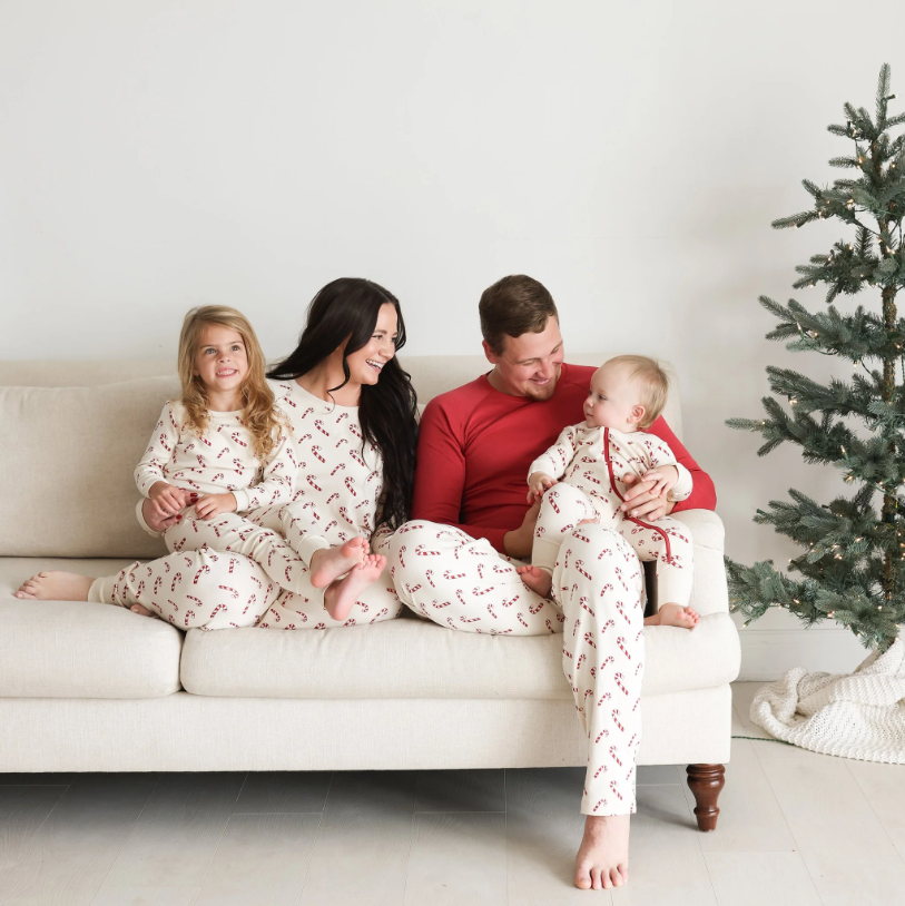 family sitting together on a couch wearing matching parade organics pajamas. Mom and big sister are wearing two piece cream colored long sleeve pajamas with a candy cane print. Baby is wearing a long sleeve onesie with the same candy cane print. Dad is wearing pants with the same candy cane print and a solid red long sleeve shirt. 